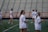 A group of women wearing white sports attire are on a sports field with lacrosse sticks. The scene suggests a practice or warm-up session in progress. In the background, there are blurred empty bleachers and a red fence.