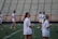 A group of women wearing white sports attire are on a sports field with lacrosse sticks. The scene suggests a practice or warm-up session in progress. In the background, there are blurred empty bleachers and a red fence.