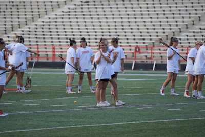 A group of women are on a sports field, wearing matching white t-shirts and shorts. They are holding lacrosse sticks and appear to be preparing for a practice or game. The field is surrounded by empty bleachers and a red railing.
