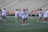 A group of women are on a sports field, wearing matching white t-shirts and shorts. They are holding lacrosse sticks and appear to be preparing for a practice or game. The field is surrounded by empty bleachers and a red railing.