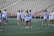 A group of women are on a sports field, wearing matching white t-shirts and shorts. They are holding lacrosse sticks and appear to be preparing for a practice or game. The field is surrounded by empty bleachers and a red railing.