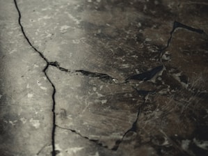 A close-up view of a cracked and weathered concrete surface with visible fractures and dark lines running through it.