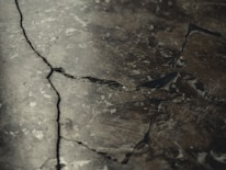 A close-up view of a cracked and weathered concrete surface with visible fractures and dark lines running through it.