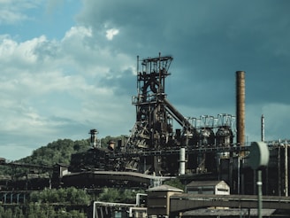A large industrial complex with towering chimneys and metal structures, set against a backdrop of lush green hills and a cloudy sky.