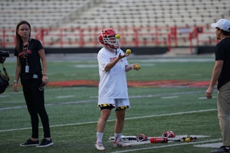 A lacrosse player is juggling three lacrosse balls on a field. The player is wearing a helmet and a white jersey. There are two other people in the scene, one holding a camera and another standing close by. Lacrosse gear is placed on the ground near the player. The background features empty bleacher seats.