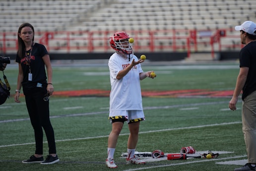 A lacrosse player is juggling three lacrosse balls on a field. The player is wearing a helmet and a white jersey. There are two other people in the scene, one holding a camera and another standing close by. Lacrosse gear is placed on the ground near the player. The background features empty bleacher seats.