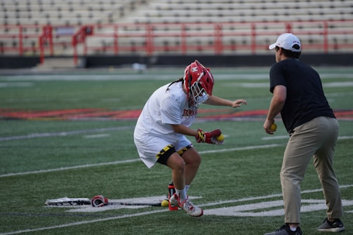 A lacrosse player in protective gear and a red helmet is practicing on a sports field. The player is holding lacrosse balls and is engaged with a coach or instructor. The setting includes stadium seating and red fencing in the background.
