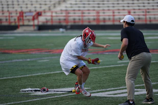 A lacrosse player in protective gear and a red helmet is practicing on a sports field. The player is holding lacrosse balls and is engaged with a coach or instructor. The setting includes stadium seating and red fencing in the background.