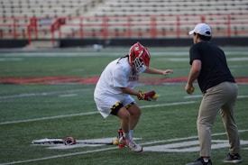 A lacrosse player in protective gear and a red helmet is practicing on a sports field. The player is holding lacrosse balls and is engaged with a coach or instructor. The setting includes stadium seating and red fencing in the background.