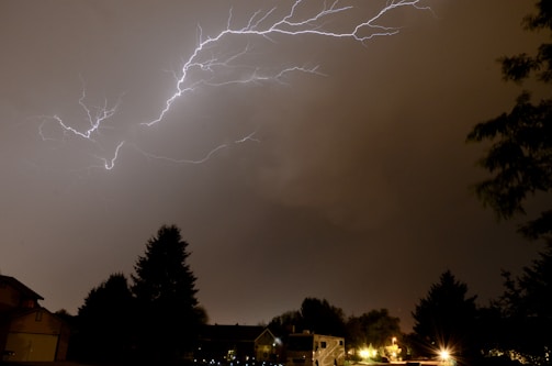 Bright lightning bolts stretch across a dark, cloudy sky over a residential area. Trees and rooftops are silhouetted against the illuminated sky, and a few artificial lights from homes and streetlights dot the landscape.