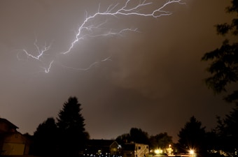 Bright lightning bolts stretch across a dark, cloudy sky over a residential area. Trees and rooftops are silhouetted against the illuminated sky, and a few artificial lights from homes and streetlights dot the landscape.