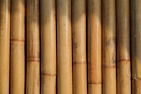 A close-up view of vertically aligned, natural bamboo stalks. The bamboo displays varying shades of light brown and tan, featuring some natural blemishes and knots. Sunlight creates shadows and highlights on the surface, emphasizing the texture and natural grains.