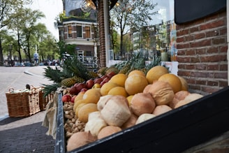 A street market display with an arrangement of fresh produce including melons, pineapples, apples, and various types of bread. The stand is located on a cobblestone street with a brick building in the background. Lush green trees and a building with classic architecture are visible across the street.