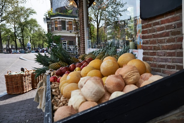 A street market display with an arrangement of fresh produce including melons, pineapples, apples, and various types of bread. The stand is located on a cobblestone street with a brick building in the background. Lush green trees and a building with classic architecture are visible across the street.