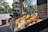 A street market display with an arrangement of fresh produce including melons, pineapples, apples, and various types of bread. The stand is located on a cobblestone street with a brick building in the background. Lush green trees and a building with classic architecture are visible across the street.