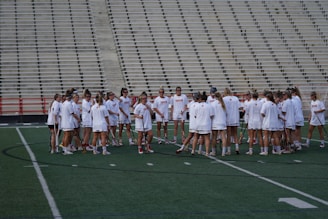 A group of athletes in white sports uniforms gather in a huddle on a green field. The backdrop features empty bleachers, and the athletes hold lacrosse sticks. The scene suggests preparation or strategy discussion before a game or practice.