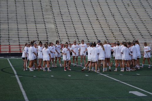 A group of athletes in white sports uniforms gather in a huddle on a green field. The backdrop features empty bleachers, and the athletes hold lacrosse sticks. The scene suggests preparation or strategy discussion before a game or practice.