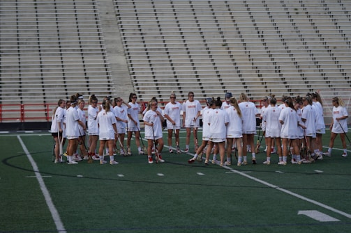 A group of athletes in white sports uniforms gather in a huddle on a green field. The backdrop features empty bleachers, and the athletes hold lacrosse sticks. The scene suggests preparation or strategy discussion before a game or practice.