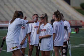 A group of young women wearing white sports jerseys stand on a lacrosse field. One of them appears to be instructing or talking to the others while holding a lacrosse stick. The background shows an empty bleacher section.