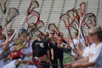 A group of people is engaged in a lacrosse activity on a sports field. Several individuals hold lacrosse sticks up high, forming an arch. The focus is on a few people in the background wearing black shirts with sunglasses, while the others wear white jerseys. The scene suggests a team or sporting event, possibly a warm-up or huddle.