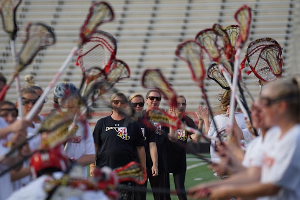 A group of people is engaged in a lacrosse activity on a sports field. Several individuals hold lacrosse sticks up high, forming an arch. The focus is on a few people in the background wearing black shirts with sunglasses, while the others wear white jerseys. The scene suggests a team or sporting event, possibly a warm-up or huddle.