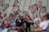 A group of people is engaged in a lacrosse activity on a sports field. Several individuals hold lacrosse sticks up high, forming an arch. The focus is on a few people in the background wearing black shirts with sunglasses, while the others wear white jerseys. The scene suggests a team or sporting event, possibly a warm-up or huddle.
