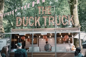 A food truck named 'The Duck Truck' is surrounded by trees in an outdoor setting. The truck has a rustic design with a wooden facade and a lit-up sign on top. Several orange lanterns hang in front and a small rubber duck is visible on the counter. People are gathered around the truck, waiting for their orders, with a hint of a festival atmosphere conveyed by the pink bunting.