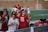 A group of athletes wearing maroon and white uniforms are gathered together on the sideline of a sports field. Some are holding water bottles and hydration equipment, while others seem engaged in conversation. There is equipment visible on a bench, including towels and bottles. The background shows a blurred view of more players or coaches and a brick building.