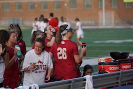 A group of athletes wearing maroon and white uniforms are gathered together on the sideline of a sports field. Some are holding water bottles and hydration equipment, while others seem engaged in conversation. There is equipment visible on a bench, including towels and bottles. The background shows a blurred view of more players or coaches and a brick building.