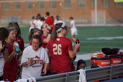 A group of athletes wearing maroon and white uniforms are gathered together on the sideline of a sports field. Some are holding water bottles and hydration equipment, while others seem engaged in conversation. There is equipment visible on a bench, including towels and bottles. The background shows a blurred view of more players or coaches and a brick building.