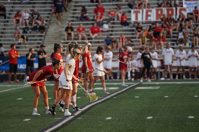 Athletes are lined up on a sports field, preparing for a game. They are dressed in athletic uniforms with some in white and others in red. The players hold lacrosse sticks and wear eye protection. In the background, spectators fill the stadium seats, mostly dressed in casual attire. A large banner is displayed behind them.