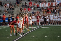 Athletes are lined up on a sports field, preparing for a game. They are dressed in athletic uniforms with some in white and others in red. The players hold lacrosse sticks and wear eye protection. In the background, spectators fill the stadium seats, mostly dressed in casual attire. A large banner is displayed behind them.