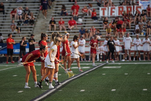 Athletes are lined up on a sports field, preparing for a game. They are dressed in athletic uniforms with some in white and others in red. The players hold lacrosse sticks and wear eye protection. In the background, spectators fill the stadium seats, mostly dressed in casual attire. A large banner is displayed behind them.