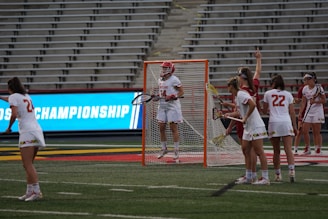 A group of female lacrosse players in white uniforms are gathered near a lacrosse goal on a sports field. One player is in front of the goal holding a stick, while others are positioned around her. The backdrop includes empty stadium bleachers and a brightly lit advertisement screen displaying 'CHAMPIONSHIP'.