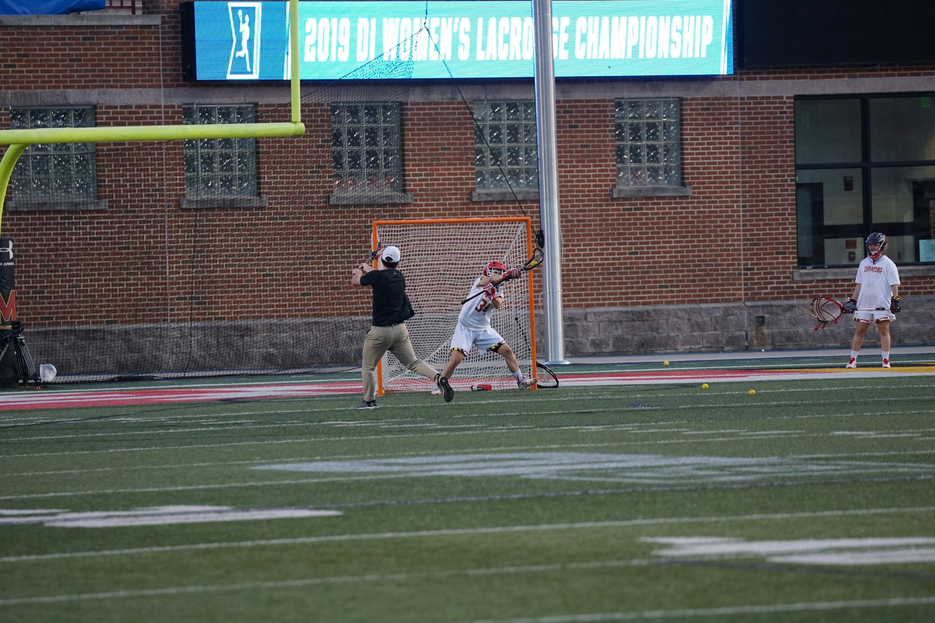 A lacrosse player in action, positioned near the goalpost, appears to be in the middle of a game or practice session. A person wearing a black shirt and beige pants holds a camera, capturing the moment. Another player stands nearby, observing. The background features a brick wall and a scoreboard displaying '2019 D1 Women's Lacrosse Championship.'