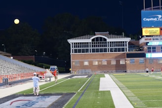 A nighttime scene featuring a sports stadium with a partially filled seating area and a large moon visible in the sky. The field is mostly empty except for a few people scattered around, and there are bright lights illuminating the venue. The stadium is surrounded by trees and a prominent sign displaying 'Maryland Lottery' is visible.