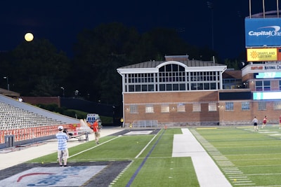 A nighttime scene featuring a sports stadium with a partially filled seating area and a large moon visible in the sky. The field is mostly empty except for a few people scattered around, and there are bright lights illuminating the venue. The stadium is surrounded by trees and a prominent sign displaying 'Maryland Lottery' is visible.
