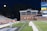 A nighttime scene featuring a sports stadium with a partially filled seating area and a large moon visible in the sky. The field is mostly empty except for a few people scattered around, and there are bright lights illuminating the venue. The stadium is surrounded by trees and a prominent sign displaying 'Maryland Lottery' is visible.