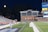 A nighttime scene featuring a sports stadium with a partially filled seating area and a large moon visible in the sky. The field is mostly empty except for a few people scattered around, and there are bright lights illuminating the venue. The stadium is surrounded by trees and a prominent sign displaying 'Maryland Lottery' is visible.