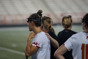 Several people wearing sports attire and protective eyewear are gathered on a sports field. They appear to be engaged in a discussion or briefing, possibly during a break from a game or practice. The background shows a blurred stadium seating, indicating an outdoor setting.