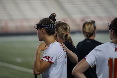 Several people wearing sports attire and protective eyewear are gathered on a sports field. They appear to be engaged in a discussion or briefing, possibly during a break from a game or practice. The background shows a blurred stadium seating, indicating an outdoor setting.