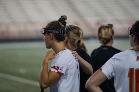 Several people wearing sports attire and protective eyewear are gathered on a sports field. They appear to be engaged in a discussion or briefing, possibly during a break from a game or practice. The background shows a blurred stadium seating, indicating an outdoor setting.