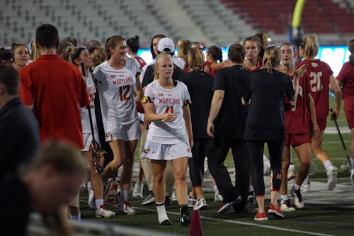 A group of female athletes on a sports field, with players wearing uniforms with 'Maryland' written on them. The scene appears to be after a match, with some players looking focused and others animatedly interacting. The setting is likely a stadium, evident by the stands in the background.