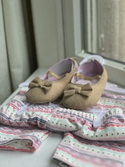 A pair of brown baby shoes with bows are placed on a patterned baby outfit with soft colors. The shoes have a soft, felt-like texture and are positioned on a hanger. The outfit features pastel hues with stripes and dotted patterns, creating a gentle and charming appearance.