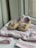 A pair of brown baby shoes with bows are placed on a patterned baby outfit with soft colors. The shoes have a soft, felt-like texture and are positioned on a hanger. The outfit features pastel hues with stripes and dotted patterns, creating a gentle and charming appearance.