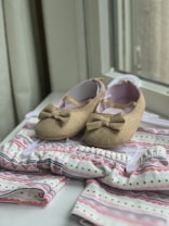A pair of brown baby shoes with bows are placed on a patterned baby outfit with soft colors. The shoes have a soft, felt-like texture and are positioned on a hanger. The outfit features pastel hues with stripes and dotted patterns, creating a gentle and charming appearance.