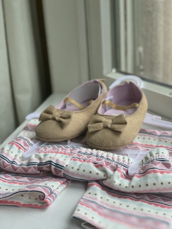 A pair of brown baby shoes with bows are placed on a patterned baby outfit with soft colors. The shoes have a soft, felt-like texture and are positioned on a hanger. The outfit features pastel hues with stripes and dotted patterns, creating a gentle and charming appearance.