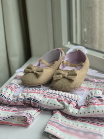 A pair of brown baby shoes with bows are placed on a patterned baby outfit with soft colors. The shoes have a soft, felt-like texture and are positioned on a hanger. The outfit features pastel hues with stripes and dotted patterns, creating a gentle and charming appearance.