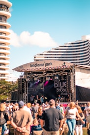 A music festival scene with a band performing on an outdoor stage. The stage is labeled 'Blues on Broadbeach Music'. A large crowd of people is gathered in front of the stage, enjoying the performance. Modern high-rise buildings are visible in the background under a clear blue sky.