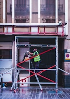 A person wearing a reflective safety vest stands on a ladder supported by a scaffold. The setting is an urban environment with a tiled floor. Behind, there is the exterior of a building with large windows reflecting intricate architectural details. Signs are present on the walls, and the ladder and scaffold assembly is secured with red crossbars.